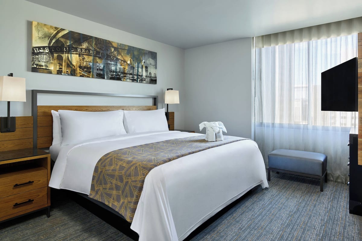 A hotel room with a large bed, two bedside tables, a bench, a TV, and a cityscape painting on the wall.