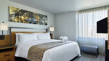 Quarto de hotel com uma cama grande, duas mesas de cabeceira, um banquinho, uma TV e uma pintura de paisagem urbana na parede.