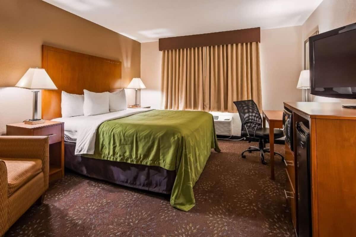 A hotel room with a bed, two bedside tables with lamps, a desk with a television, a chair, and a large window with curtains.