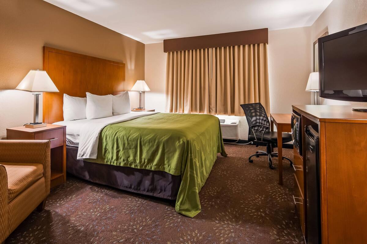 A hotel room with a bed, two bedside tables with lamps, a desk with a television, a chair, and a large window with curtains.
