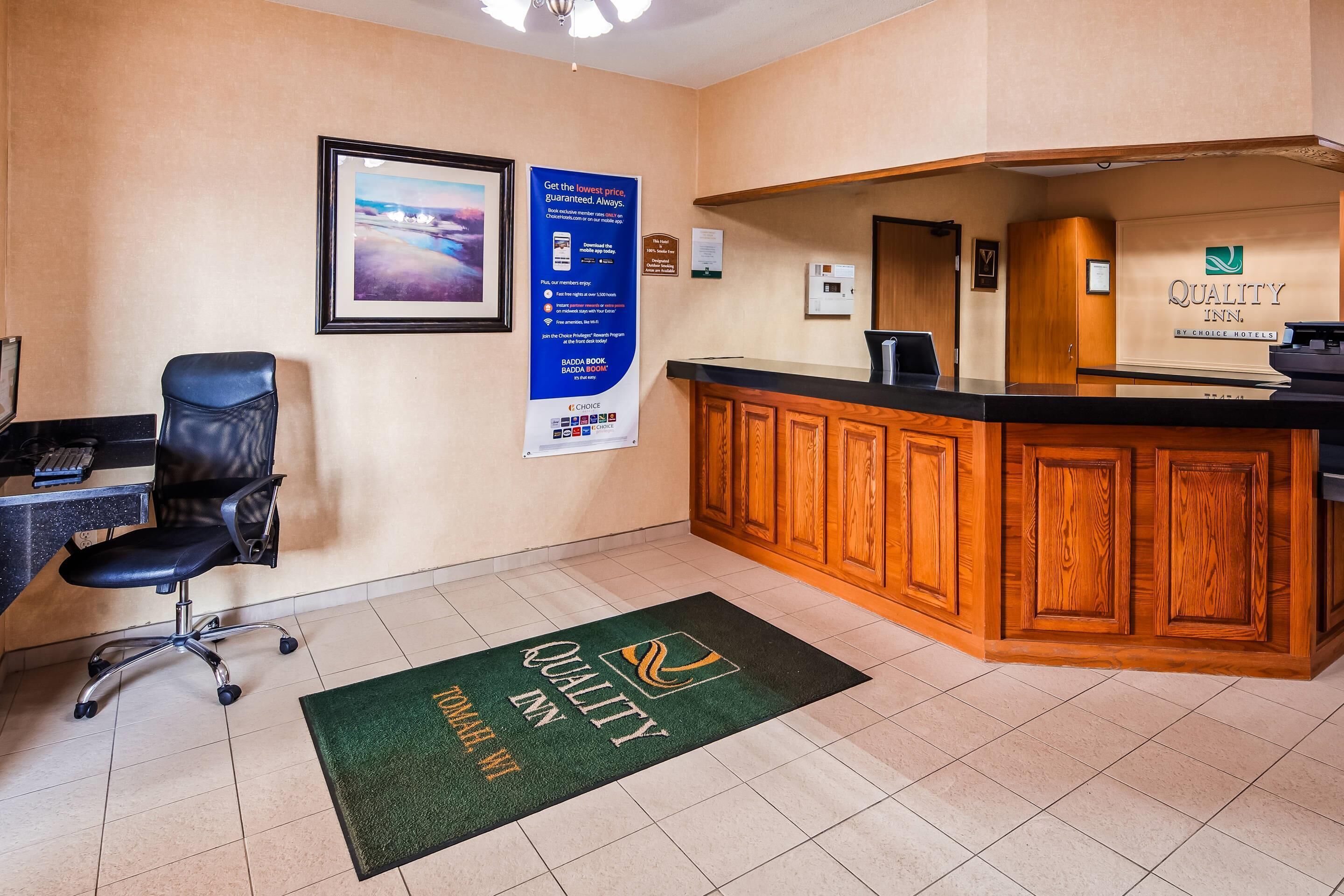 A hotel reception area with a wooden reception desk, a black office chair, a framed picture on the wall, and a green doormat with the hotel logo.