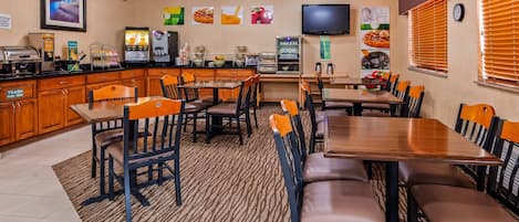 Un comedor de hotel con mesas y sillas de madera, una estación de café y un televisor en la pared.