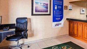A hotel reception area with a desk, a black office chair, a framed picture on the wall, a blue informational poster, and a green carpet with the hotel logo. - Quality Inn Tomah (Tomah)