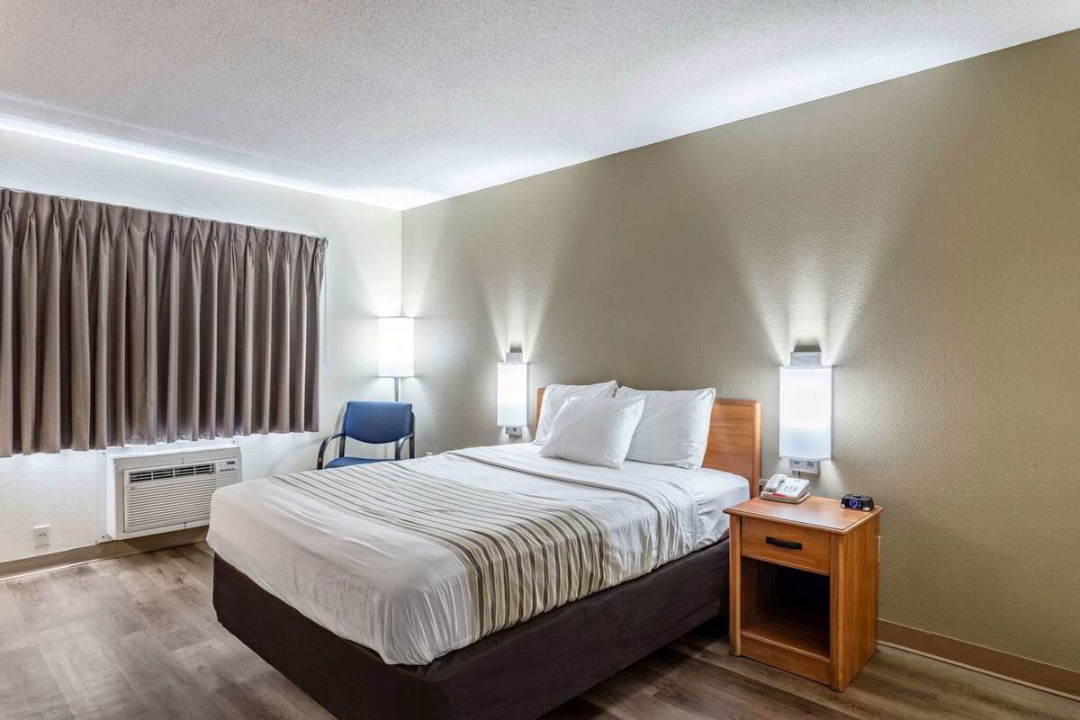Une chambre d’hôtel avec un lit, des lampes de chevet, une chaise, une table de chevet et une fenêtre avec des rideaux.