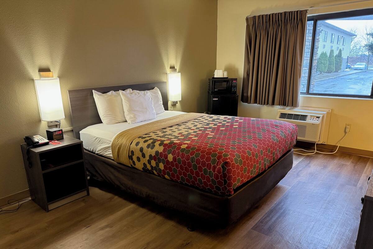 A hotel room with a bed, bedside lamps, a nightstand with a phone, a window with curtains, and a view of the outside.