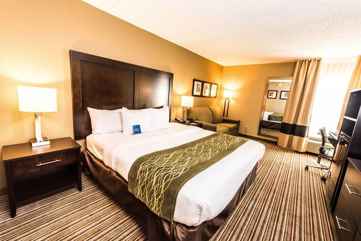 A hotel room with a large bed, a bedside table with a lamp, a chair, a desk with a computer, and a television.