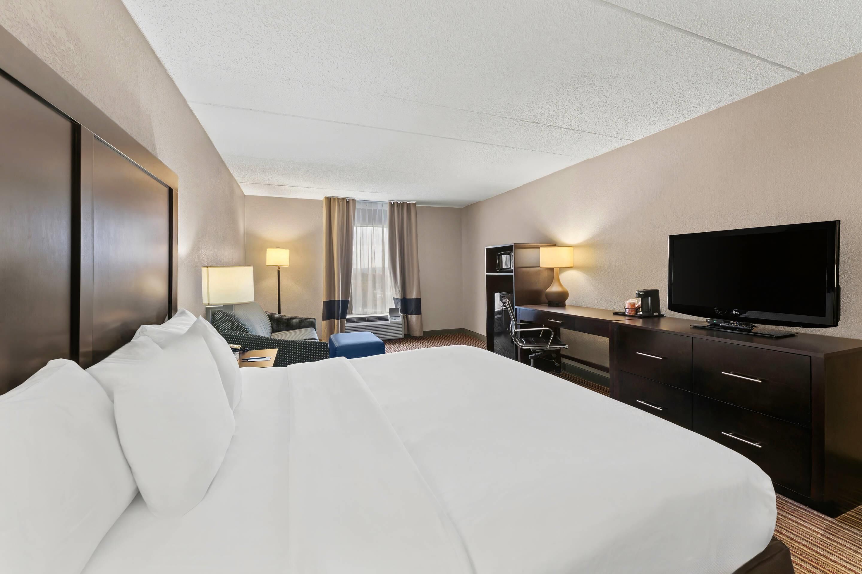 Habitación de hotel con una cama grande, un escritorio con televisor de pantalla plana, una silla, una lámpara y una cafetera.