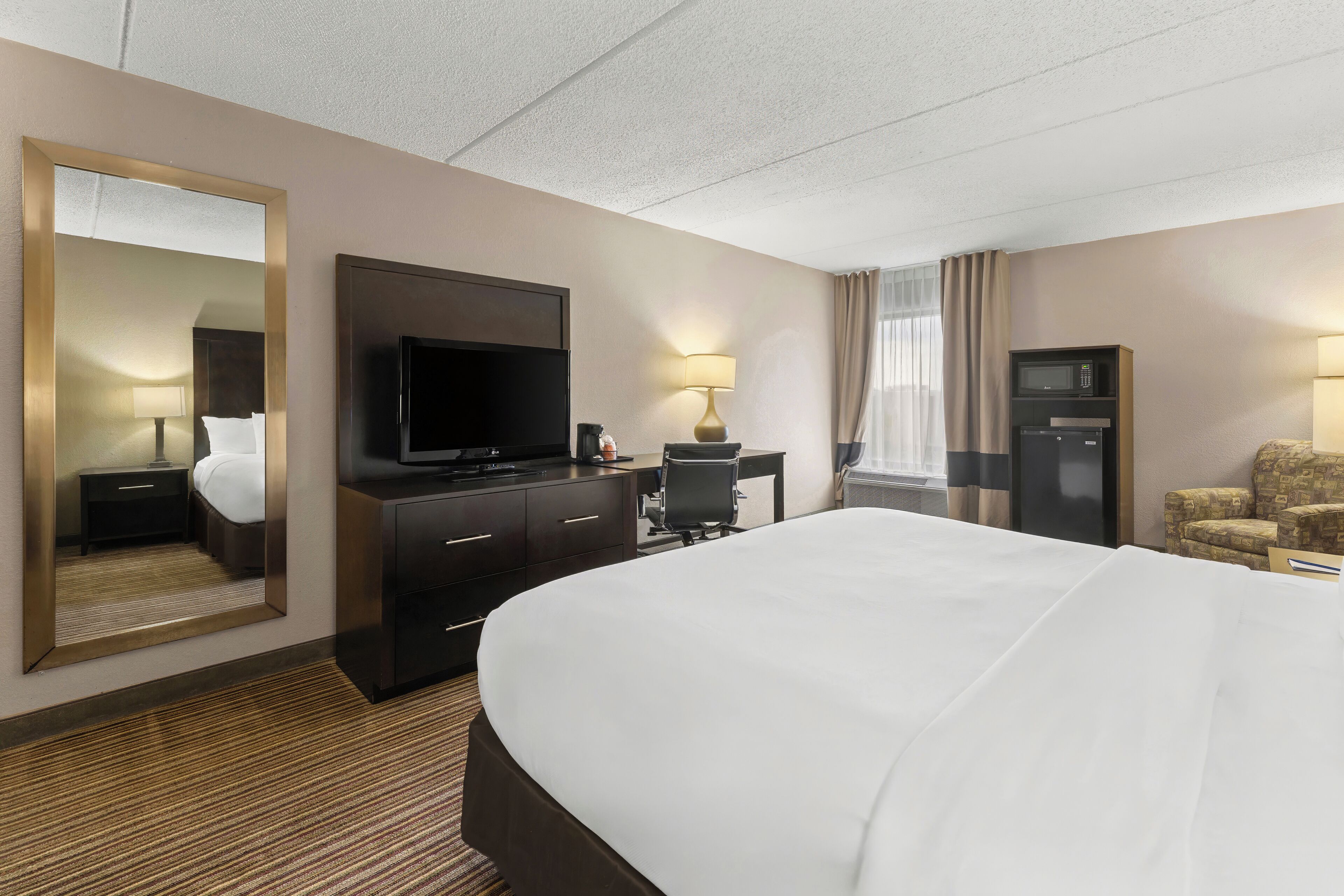 A hotel room with a large bed, a flat-screen TV, a desk with a lamp, a microwave, and a seating area with a chair.