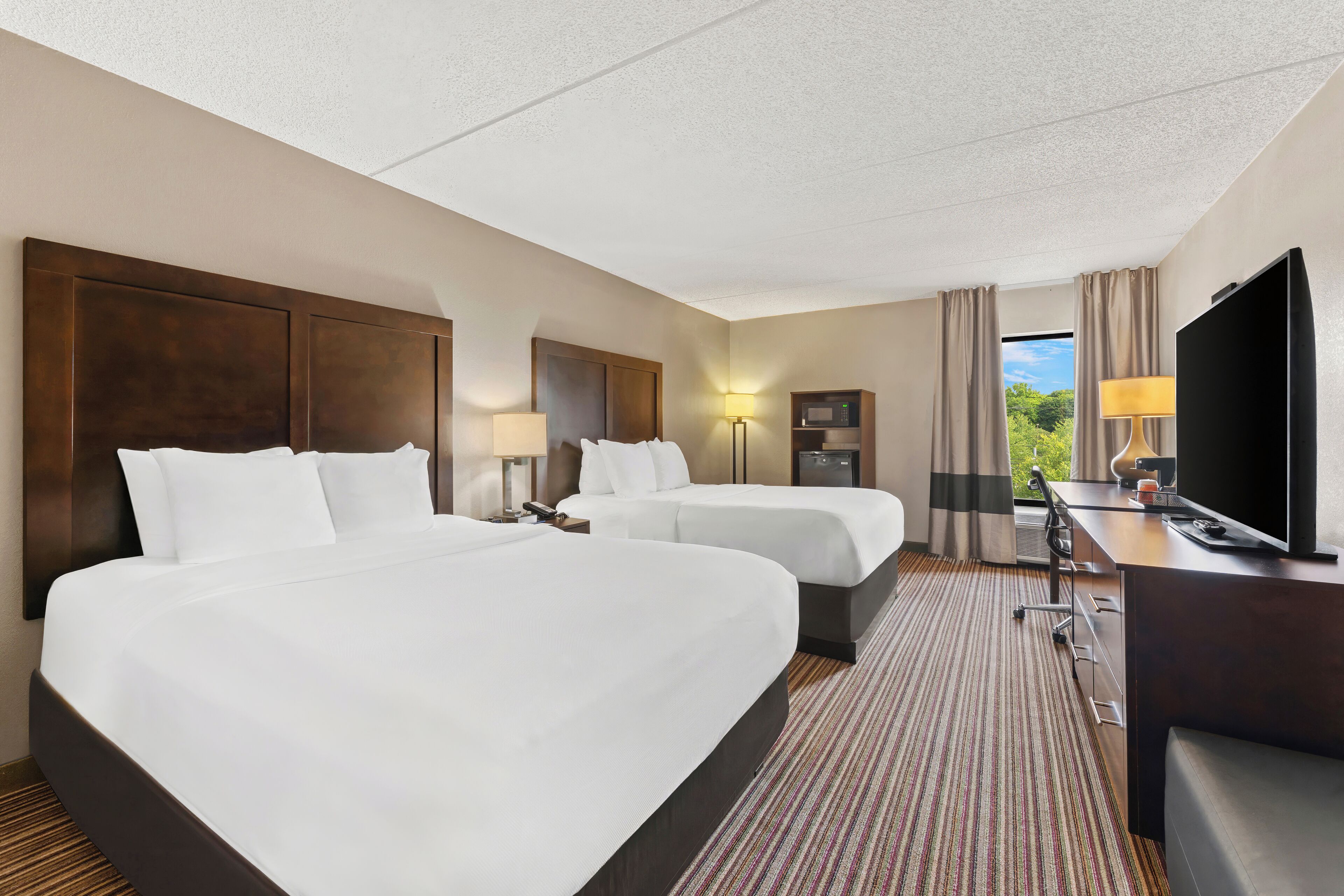 A hotel room with two beds, a desk with a TV, a lamp, and a view of greenery outside.