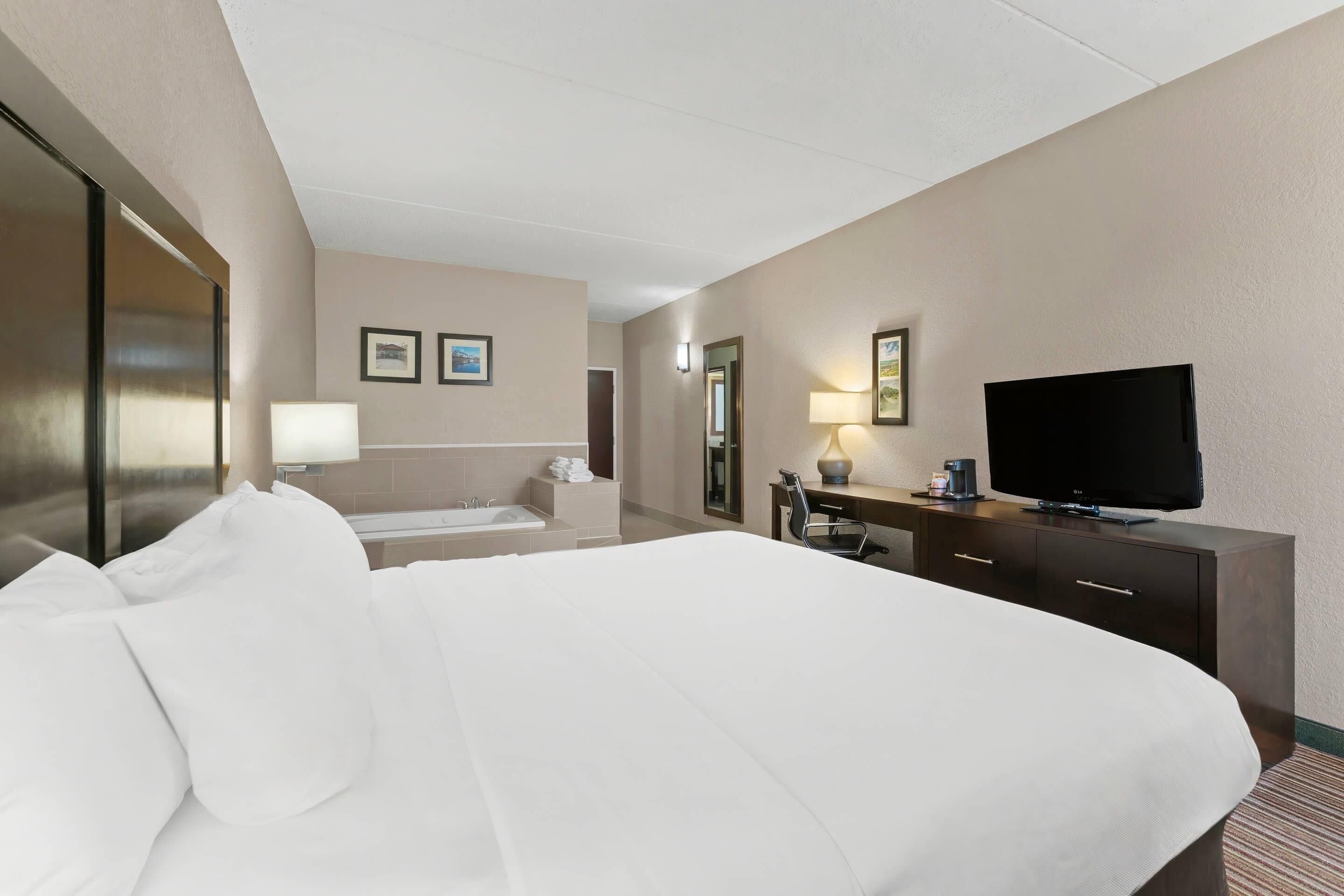 A hotel room with a large bed, a desk with a flat-screen TV, a bathroom with a bathtub, and a mirror.