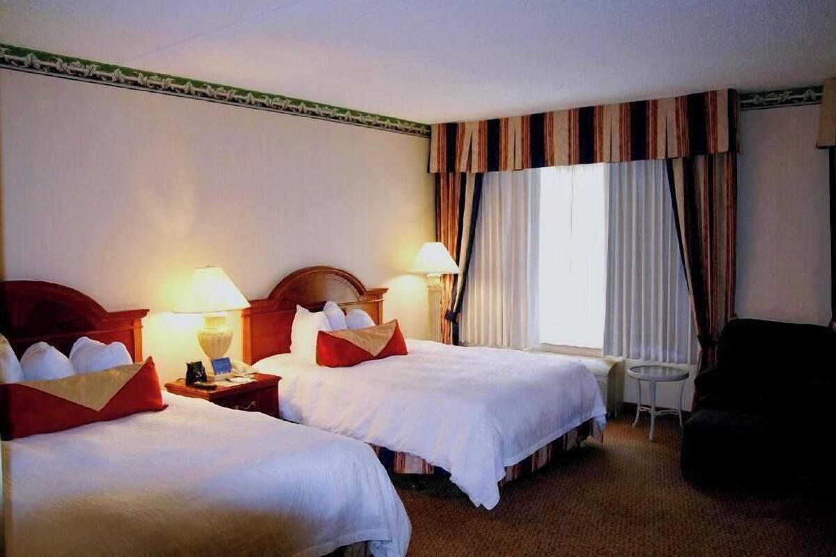 A hotel room with two beds, a chair, a small table, and a window with curtains.