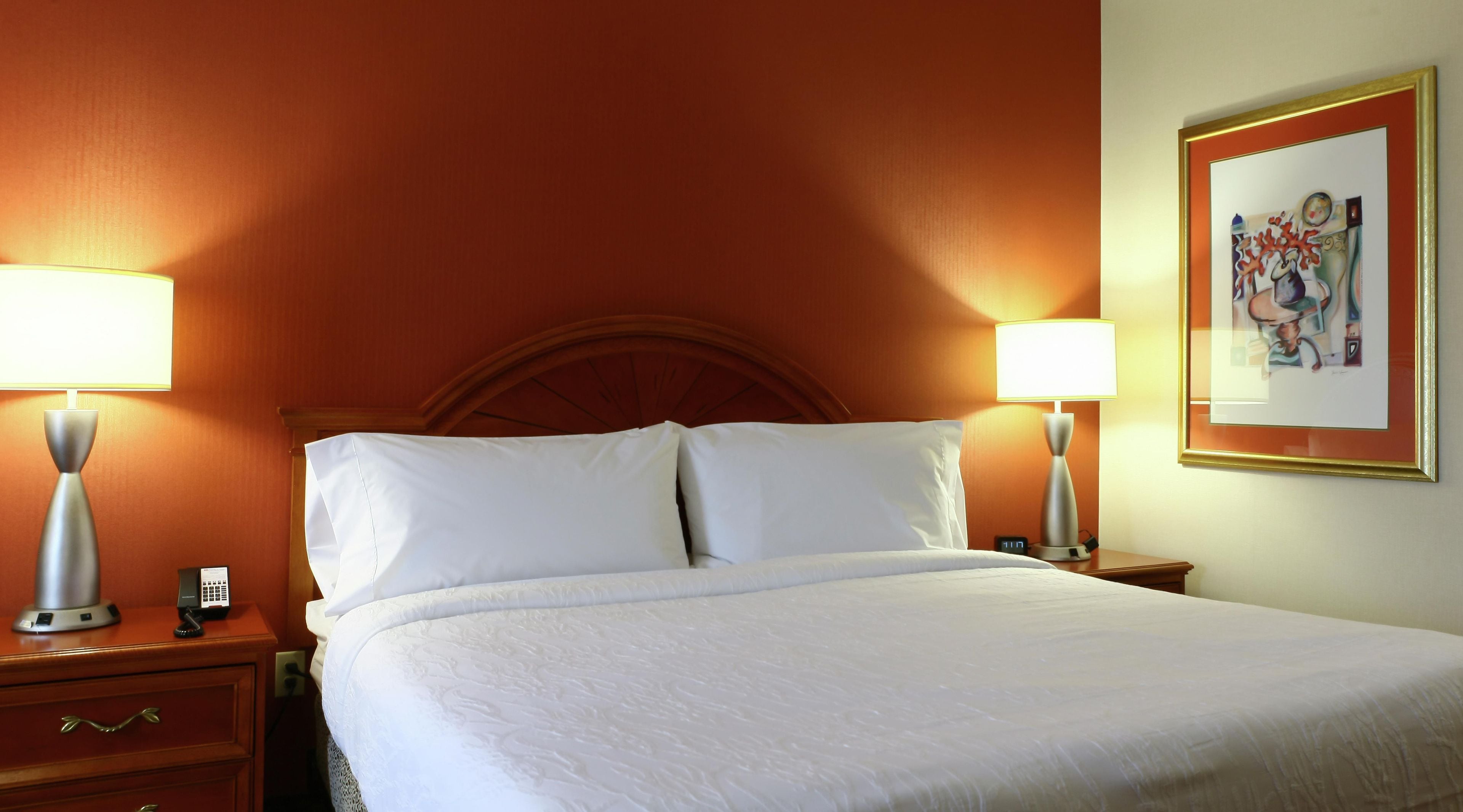 Habitación de hotel con cama, dos lámparas de noche, una mesita de noche y un cuadro en la pared.