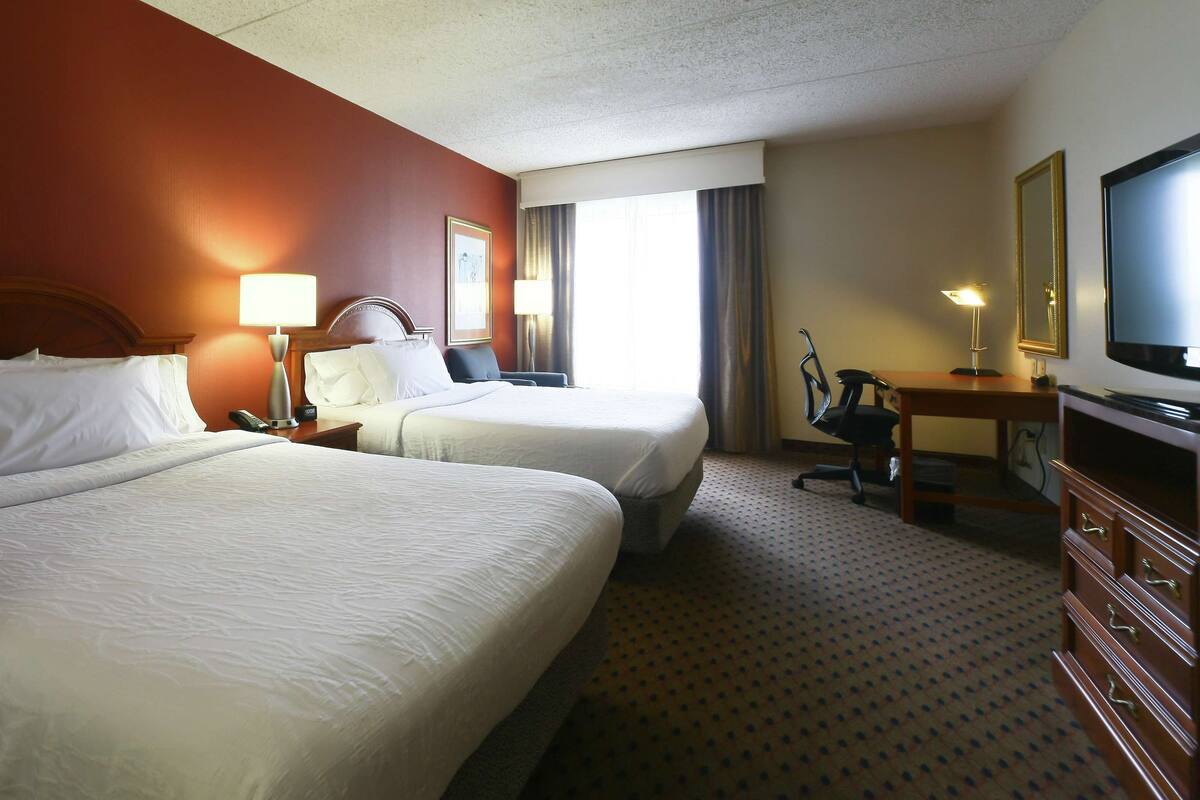 A hotel room with two beds, a desk with a chair, a television, and a window with curtains.
