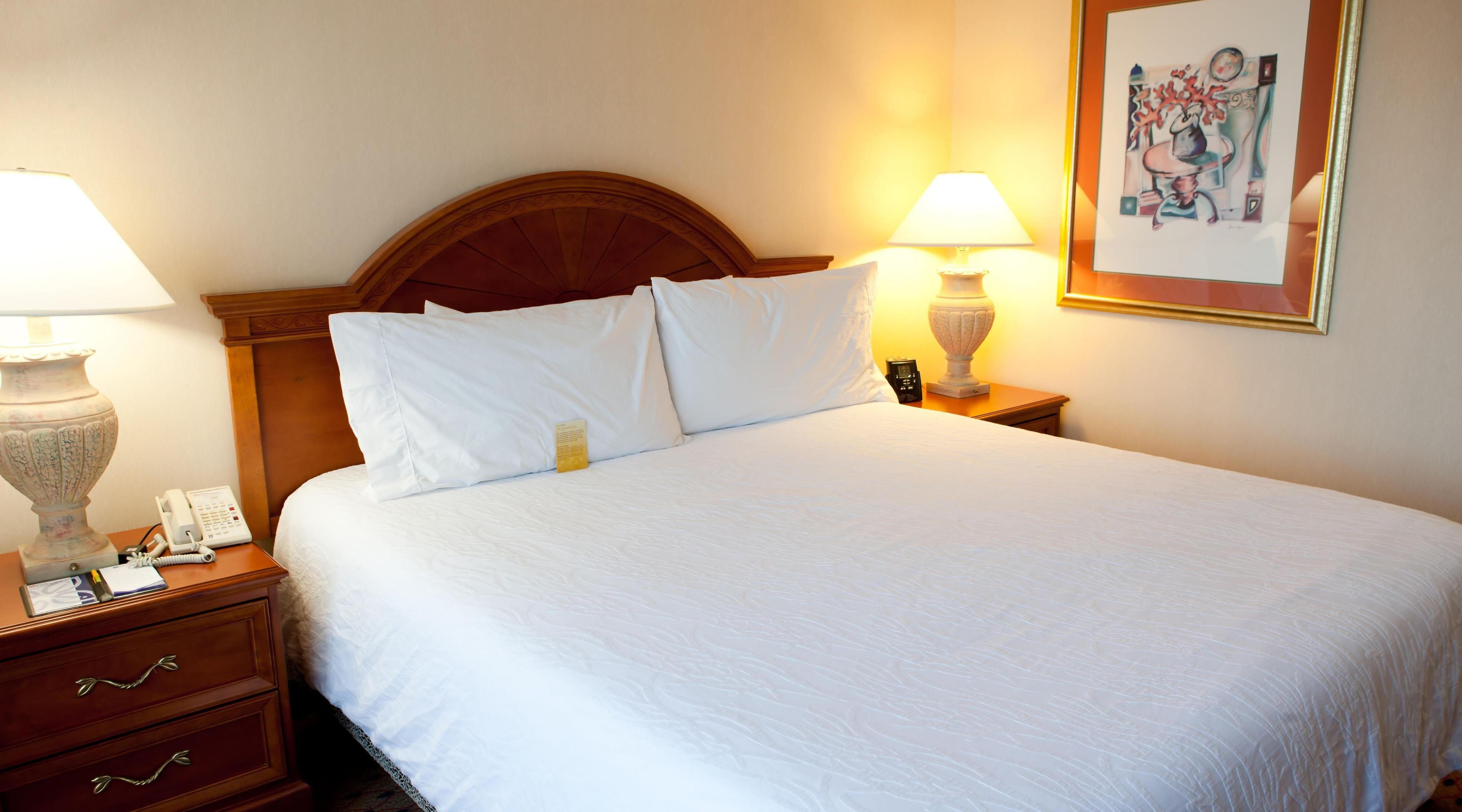 A hotel room with a large bed, two bedside tables, a nightstand, a lamp, a telephone, and a framed picture on the wall.