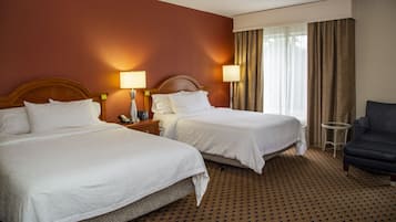 A hotel room with two beds, a nightstand with a lamp, a chair, and a window with curtains.