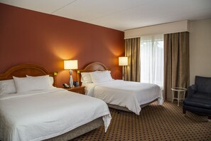 Habitación de hotel con dos camas, una mesita de noche con lámpara, una silla y una ventana con cortinas.