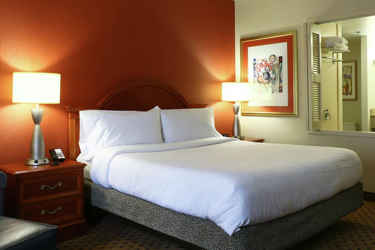 A hotel room with a large bed, two bedside lamps, a wooden nightstand, a chair, and a bathroom visible through an open door.