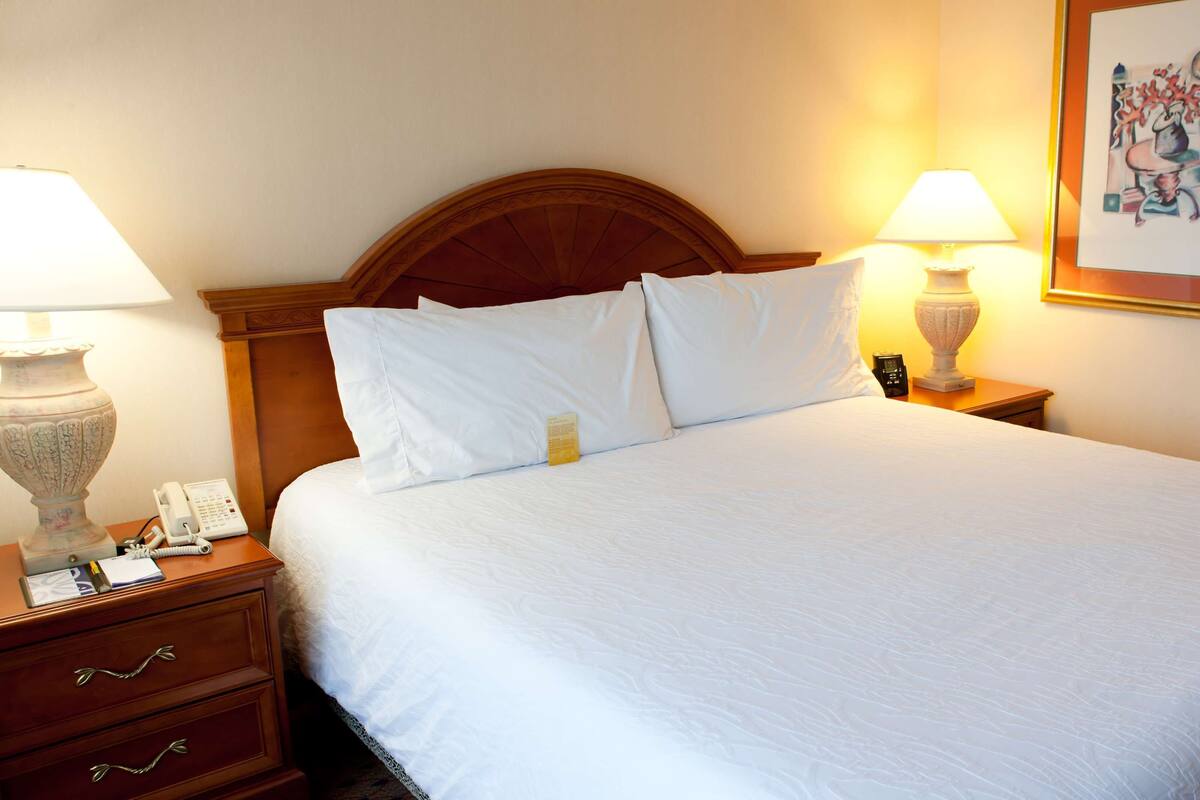 A neatly made bed with white linens, two bedside lamps, a wooden headboard, a nightstand with a telephone, and a framed picture on the wall.
