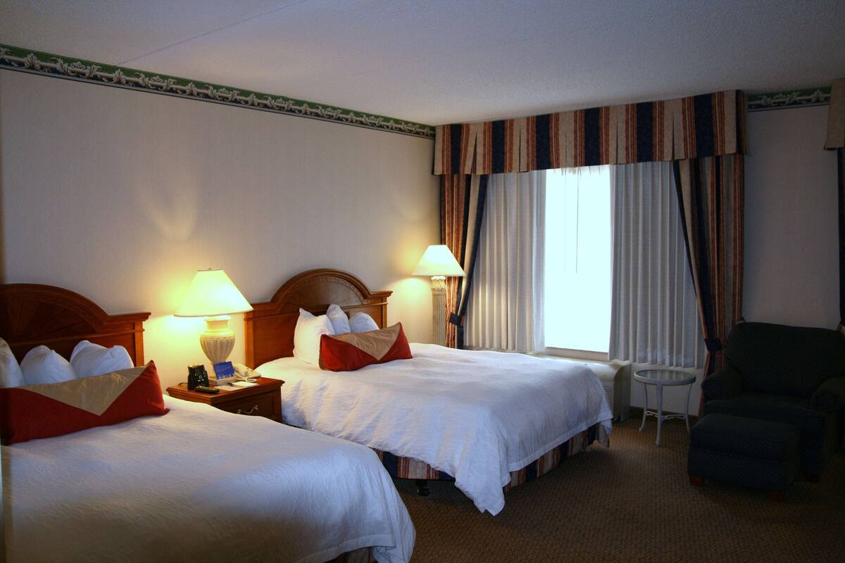 A hotel room with two beds, a small table, a chair, and a window with curtains.