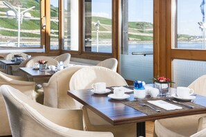 A dining area with tables set for two, chairs, and a view of the sea.
