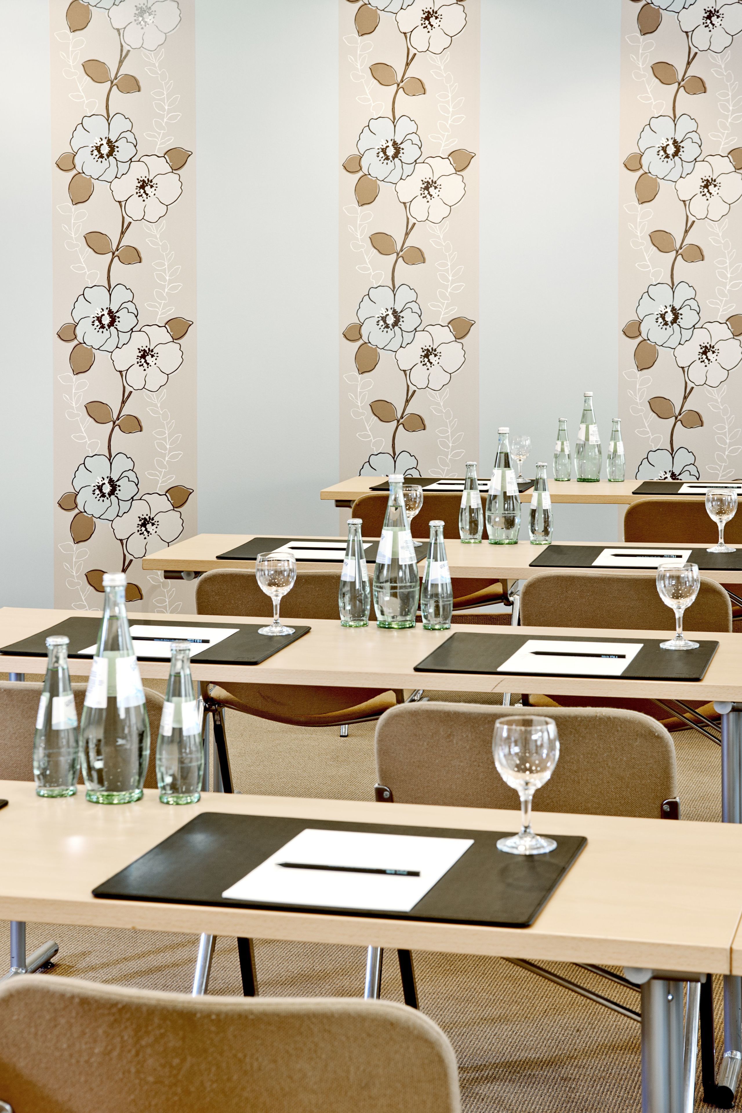 Ein Konferenzraum mit für Meetings gedeckten Tischen, ausgestattet mit Wasserflaschen und Gläsern, und mit floralem Wandpapier dekoriert.