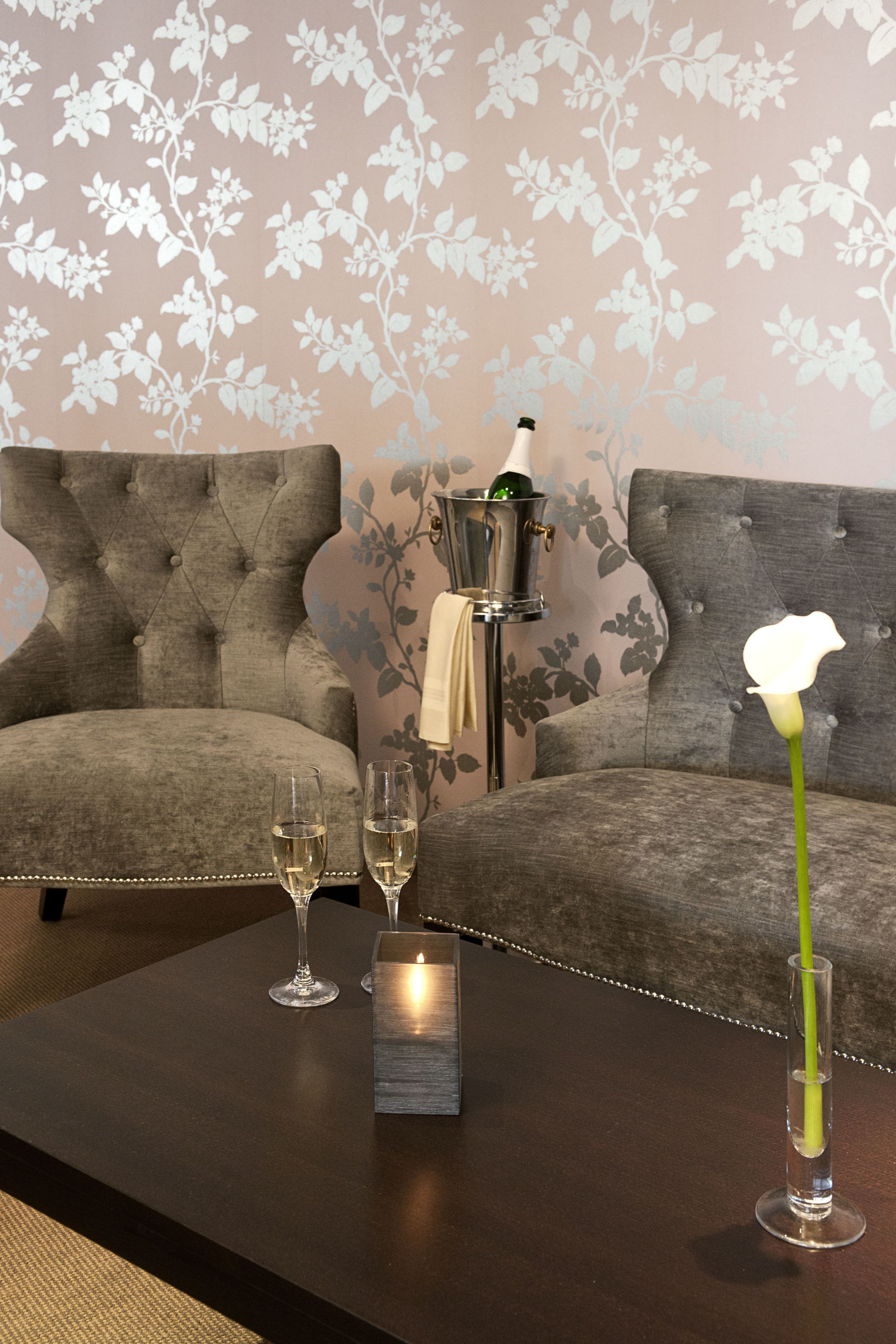 Una habitación con un sofá gris con capitoné, dos copas de vino, una vela y un florero con una flor blanca sobre una mesa de madera oscura.