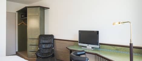 A hotel room with a bed, a desk with a computer monitor, two chairs, a wardrobe, and a radiator.