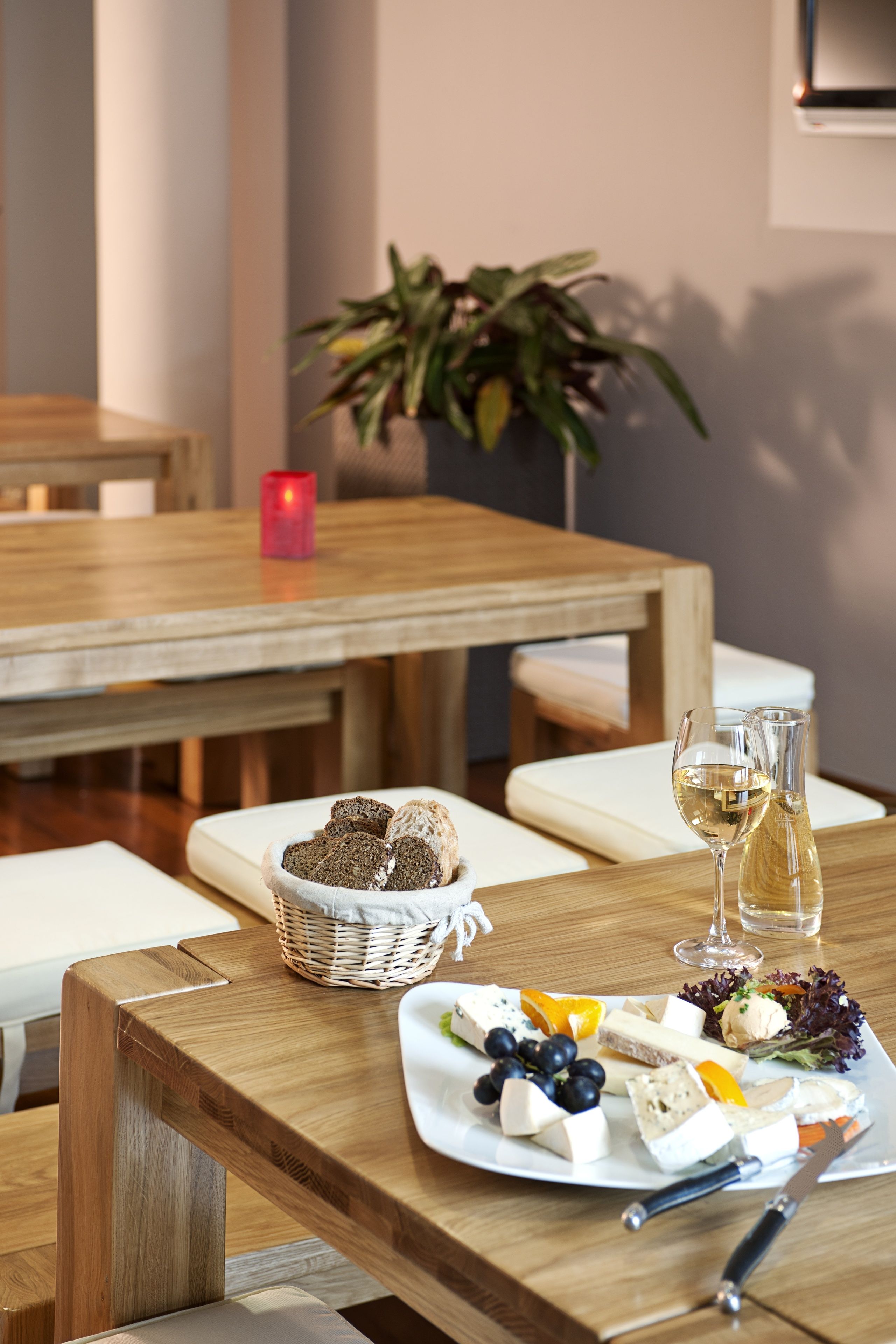 Una mesa de madera con un plato de queso, uvas y pan, una copa de vino blanco y una vela.