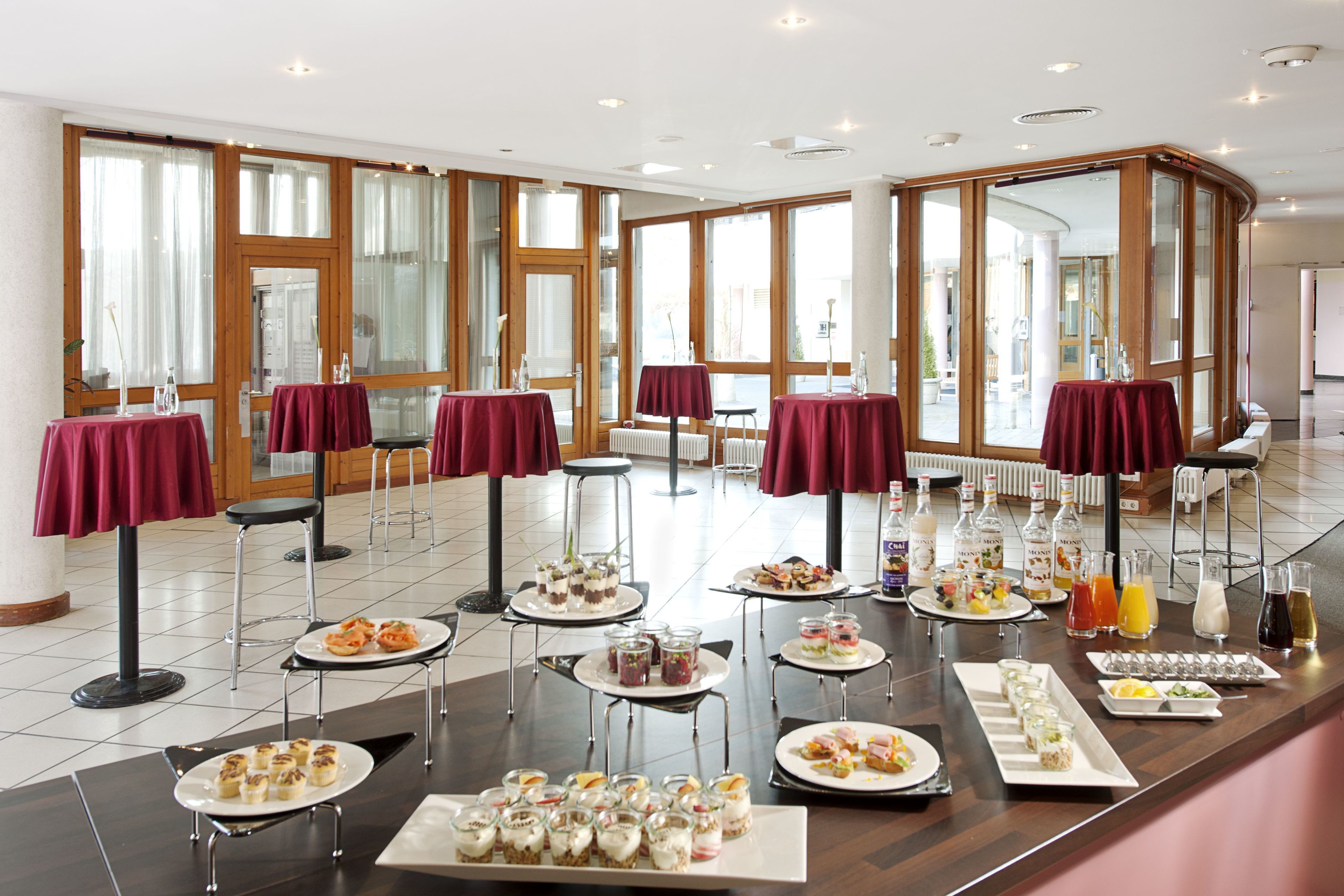 Montaje de bufé con diversos alimentos y bebidas, dispuesto en mesas con manteles rojos en una sala amplia con ventanas grandes.