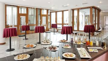 A buffet setup with various food items and drinks, arranged on tables with red tablecloths in a spacious room with large windows.
