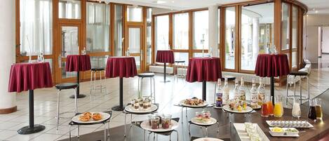 Montaje de bufé con diversos alimentos y bebidas, dispuesto en mesas con manteles rojos en una sala amplia con ventanas grandes.