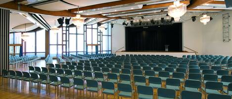 Un ampio auditorium con file di sedie verdi, travi di legno a soffitto e un palcoscenico con uno sfondo nero.