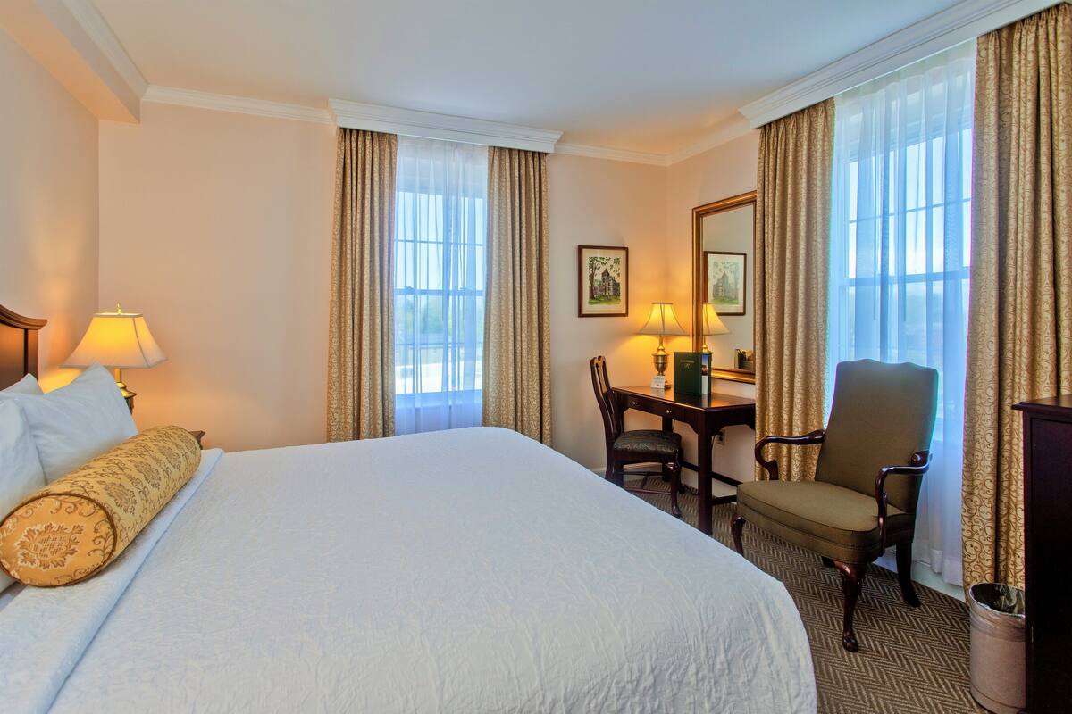 A hotel room with a large bed, a desk with a chair, a lamp, and a picture on the wall.