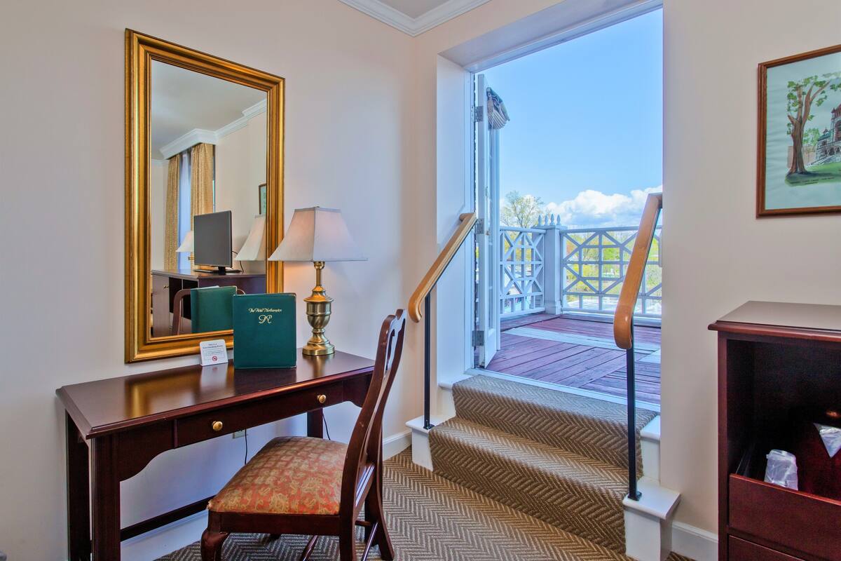 A hotel room with a desk, chair, lamp, and a large mirror. There is a staircase leading to an outdoor area with a railing.