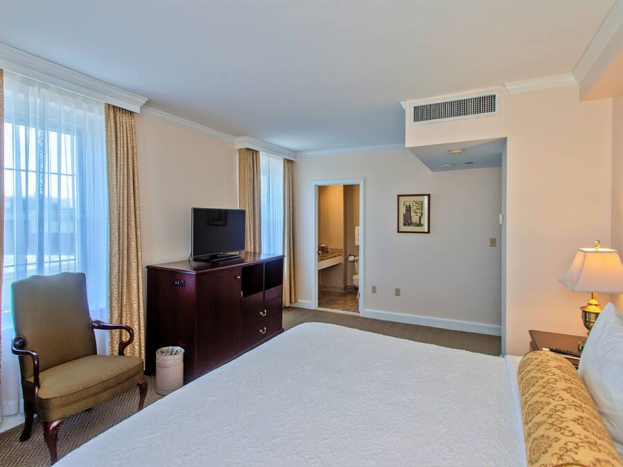 A hotel room with a large bed, a wooden chair, a desk with a television, and a bathroom visible through an open door.
