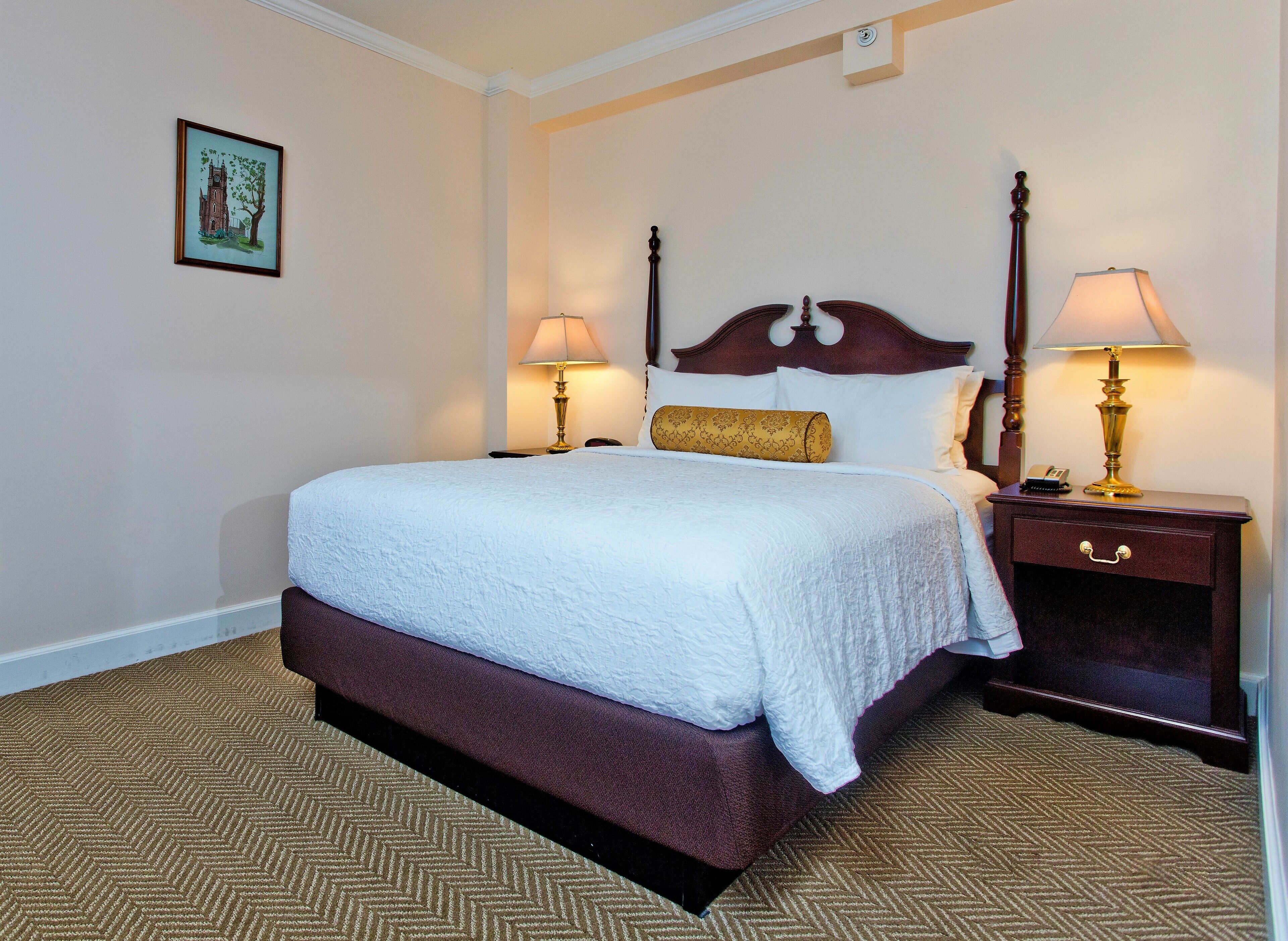 A neatly made bed with a white comforter and a brown headboard, flanked by two bedside tables with lamps, one of which has a phone and a hairbrush on it.