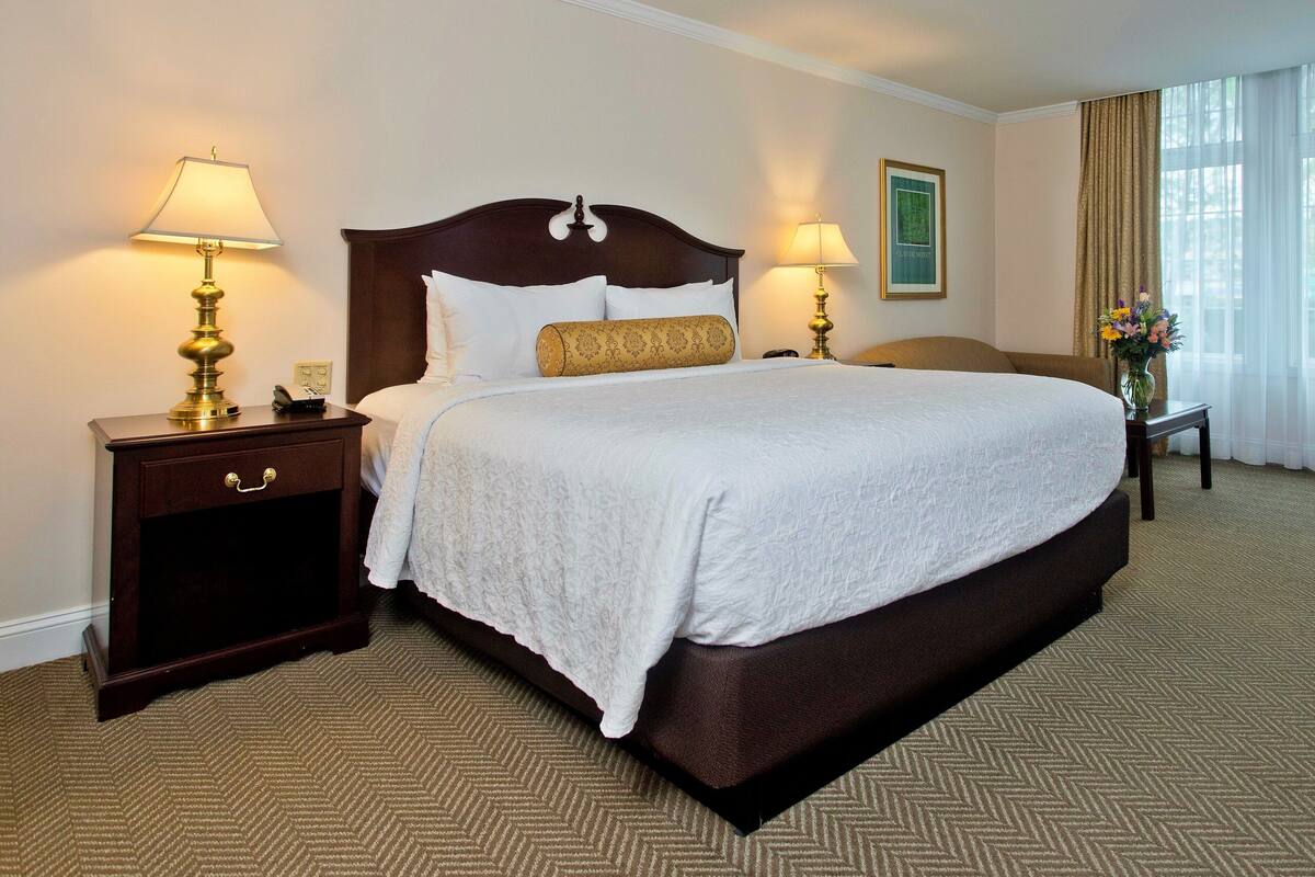 A hotel room with a large bed, two bedside tables with lamps, a nightstand with a drawer, a vase with flowers, and a window with curtains.