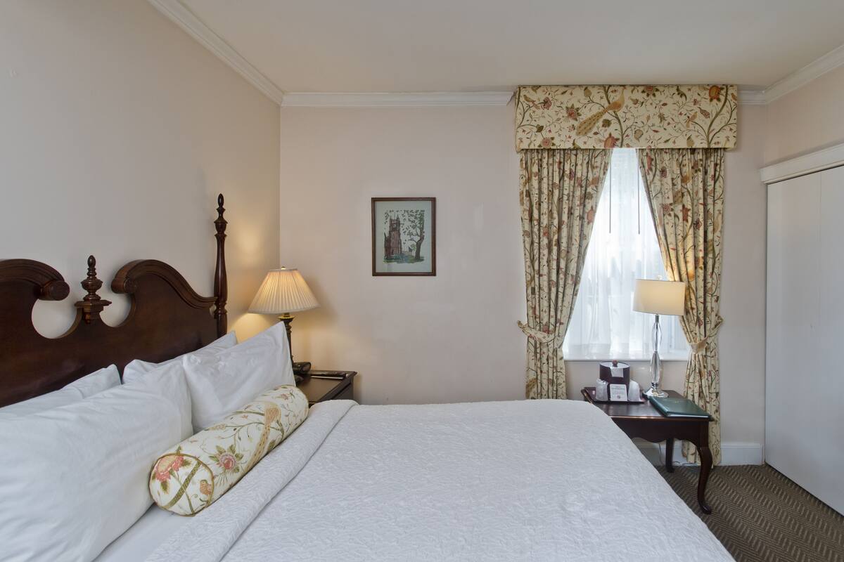 A neatly made bed with white linens and a floral pillow, a wooden headboard, a bedside table with a lamp, and a window with floral curtains.