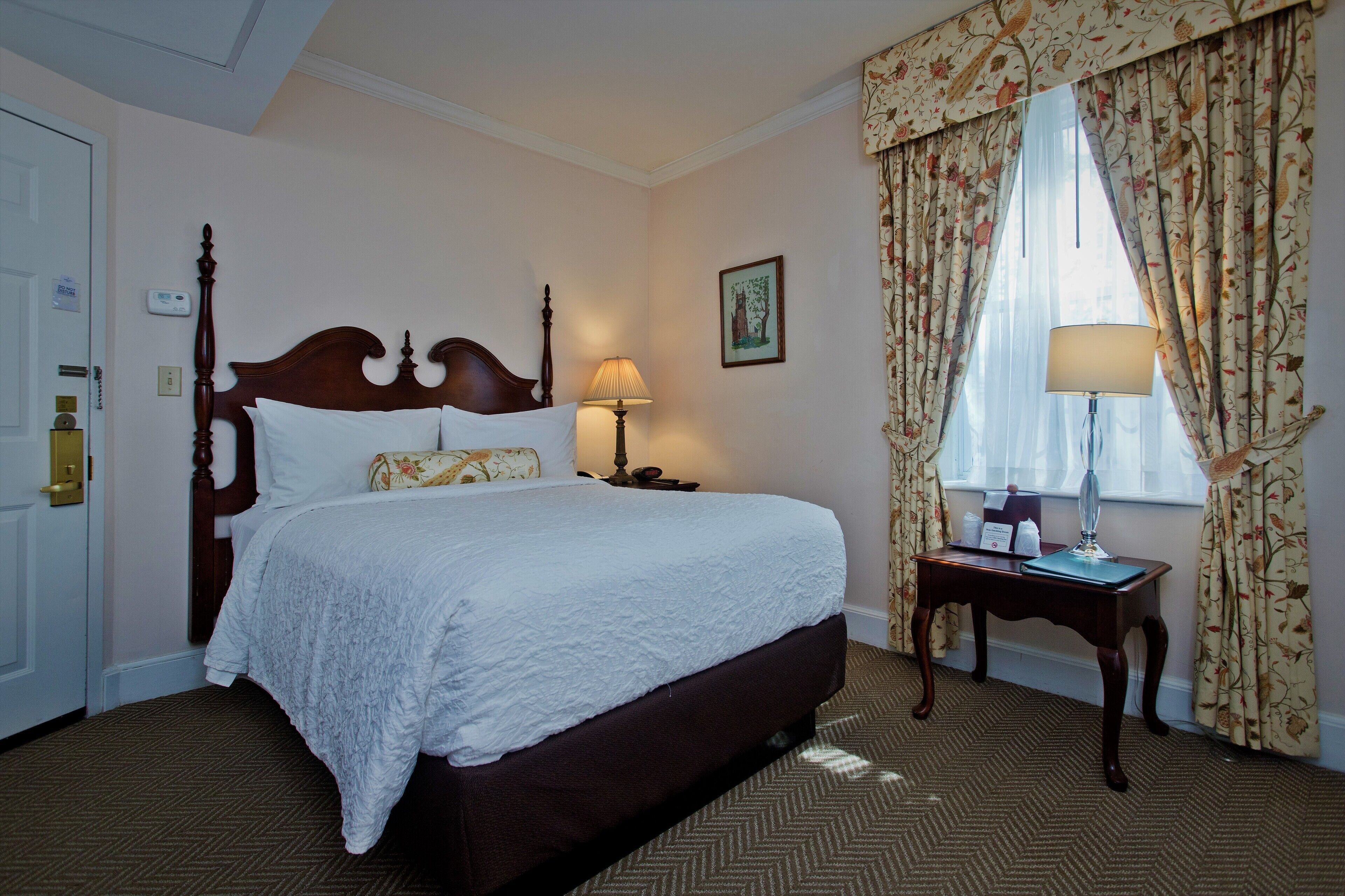 A neatly made bed with a wooden headboard, a bedside table with a lamp, and floral curtains.