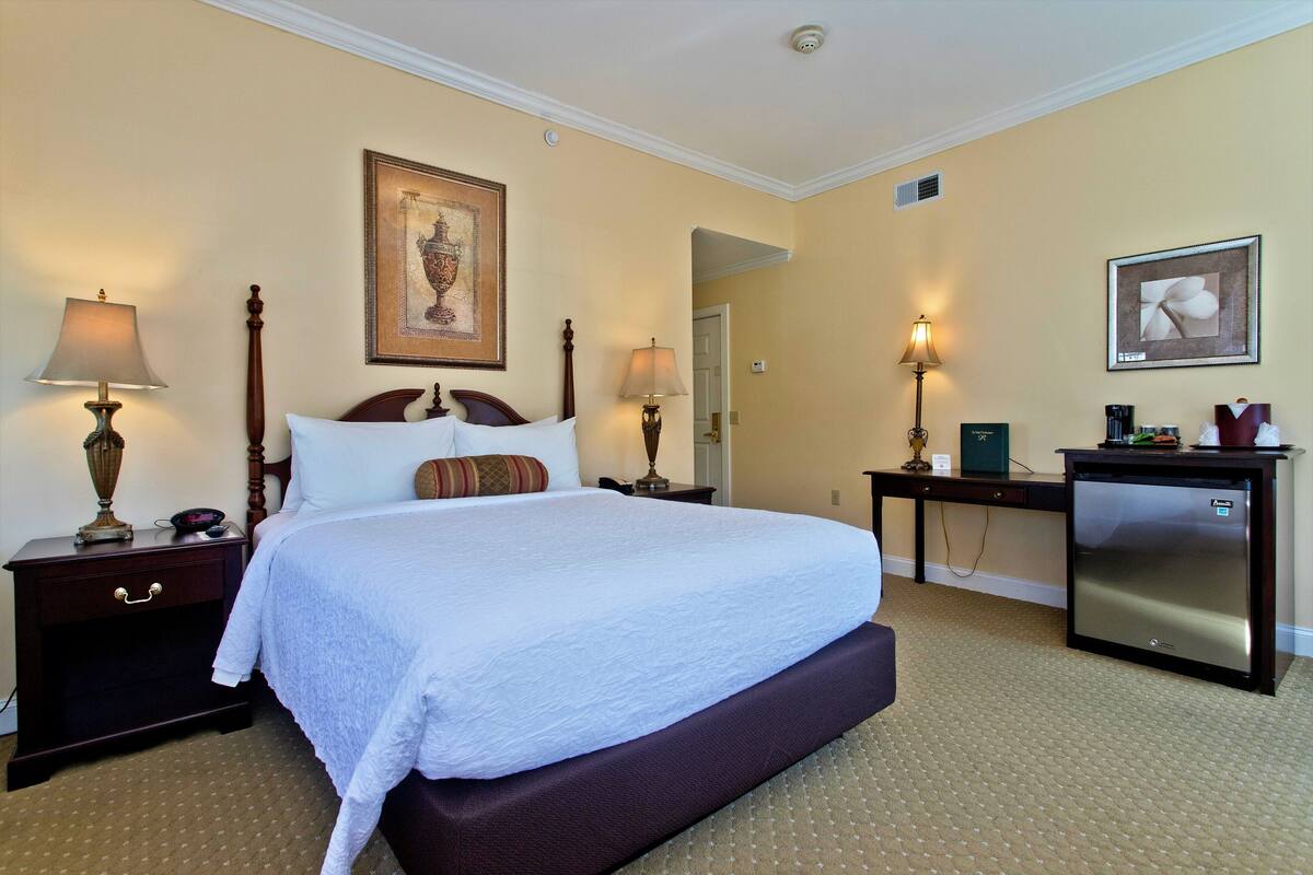 A hotel room with a large bed, two bedside tables with lamps, a desk with a refrigerator, and two framed pictures on the walls.