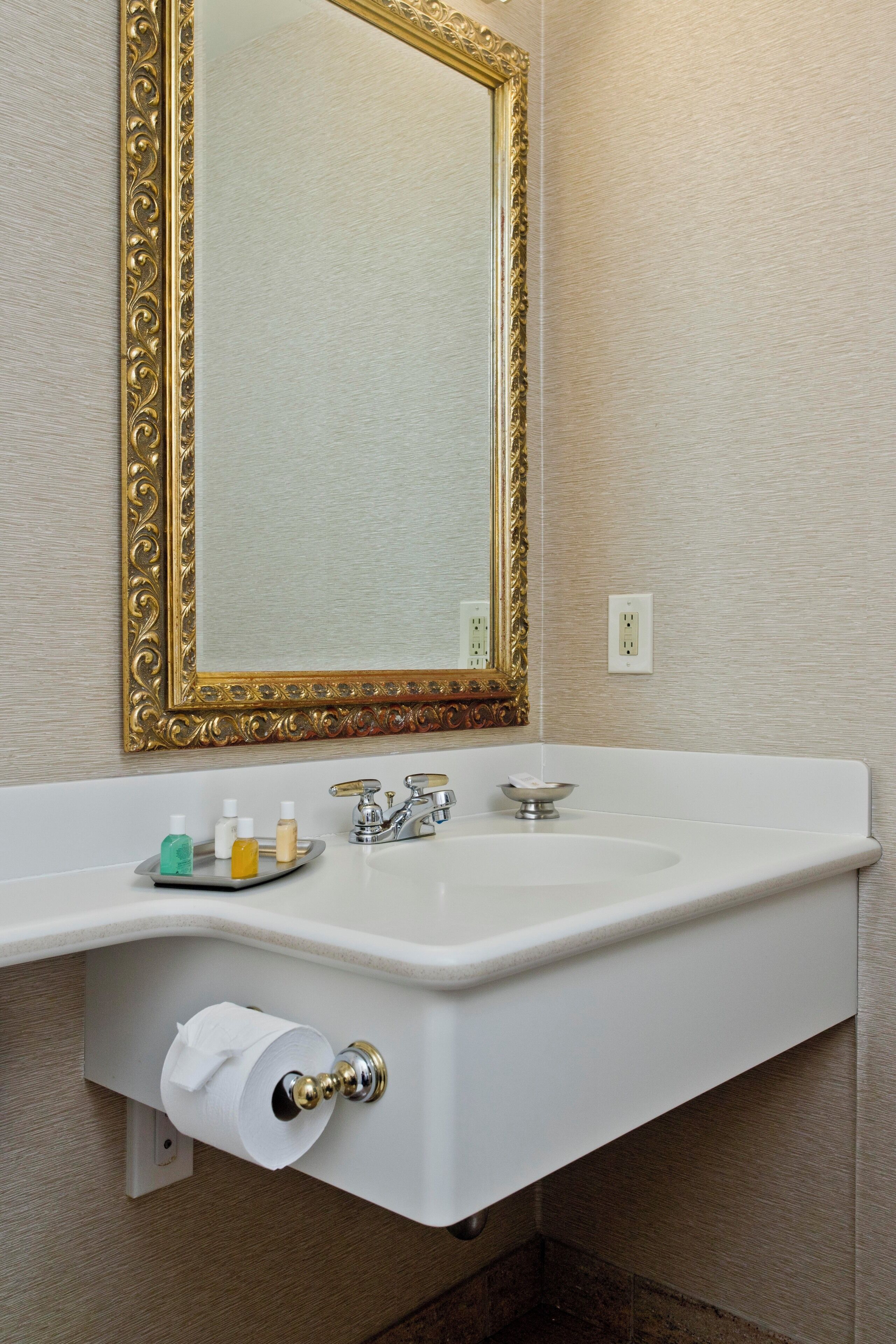 A bathroom with a white sink, gold-framed mirror, and toiletries on the counter.