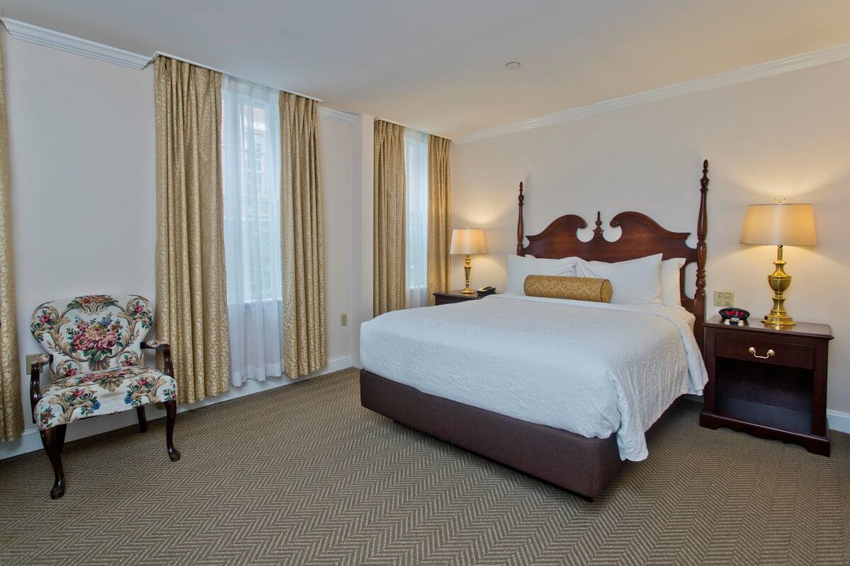 A hotel room with a large bed, two bedside tables with lamps, a floral armchair, and a herringbone-patterned carpet.