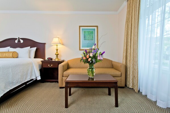 Habitación de hotel con cama, sofá, mesita de noche con lámpara, florero con flores y un cuadro en la pared.