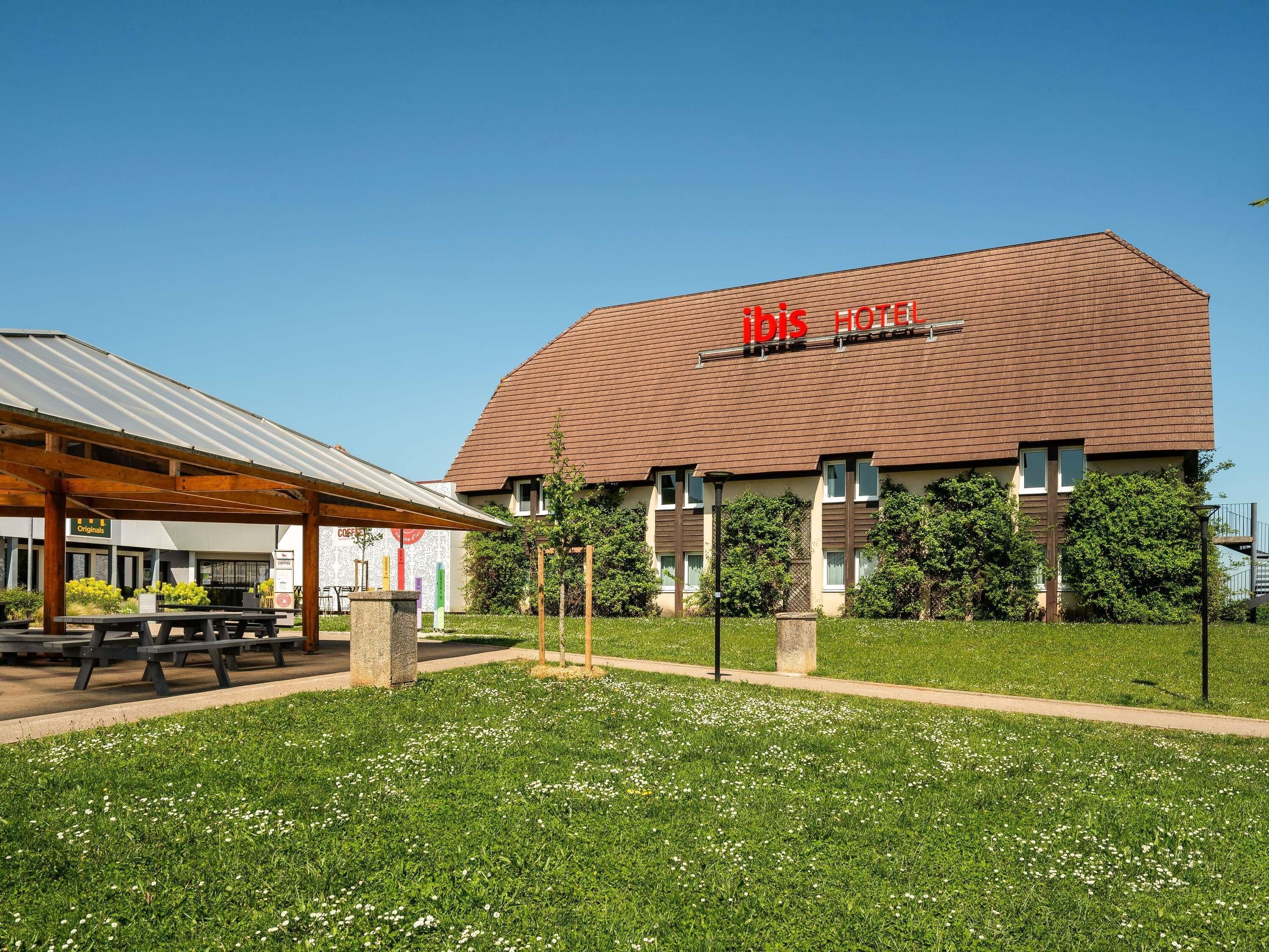 A hotel with a red sign, a covered outdoor seating area, and a well-maintained lawn.