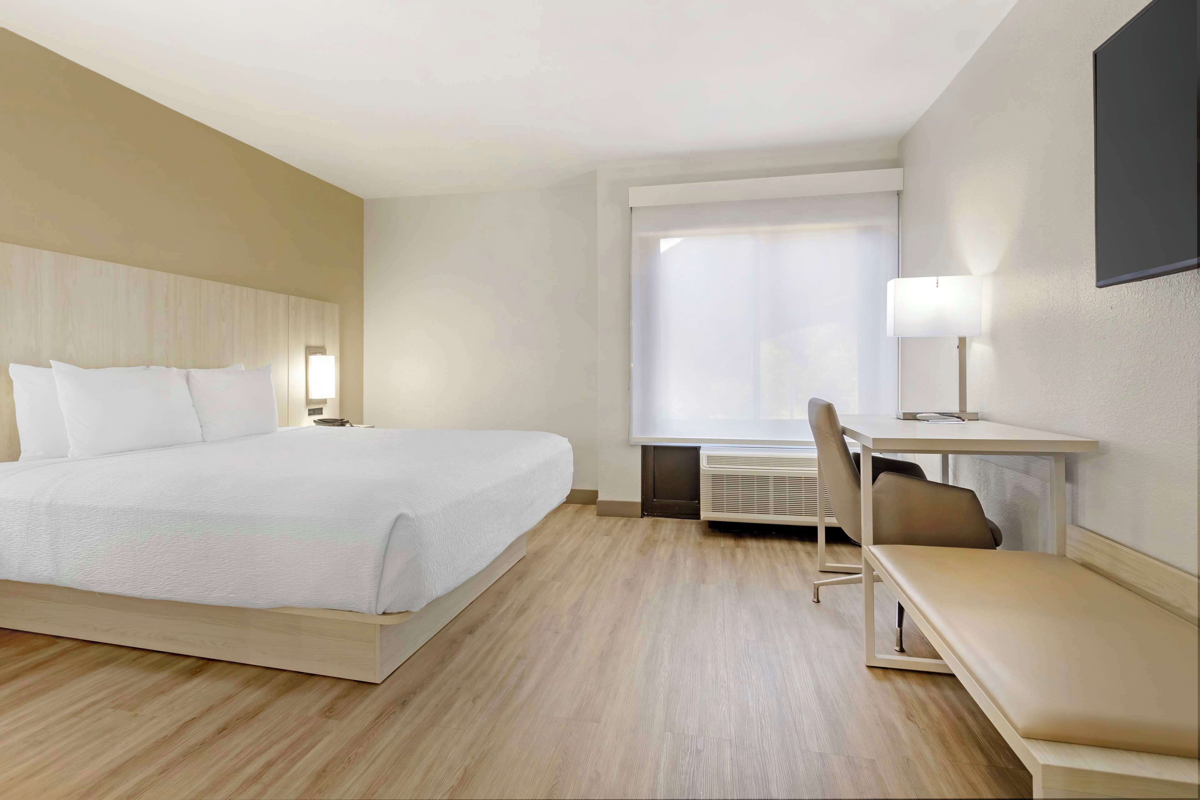 Una habitación de hotel con una cama, un escritorio, una silla, un banco y un ventanal grande.