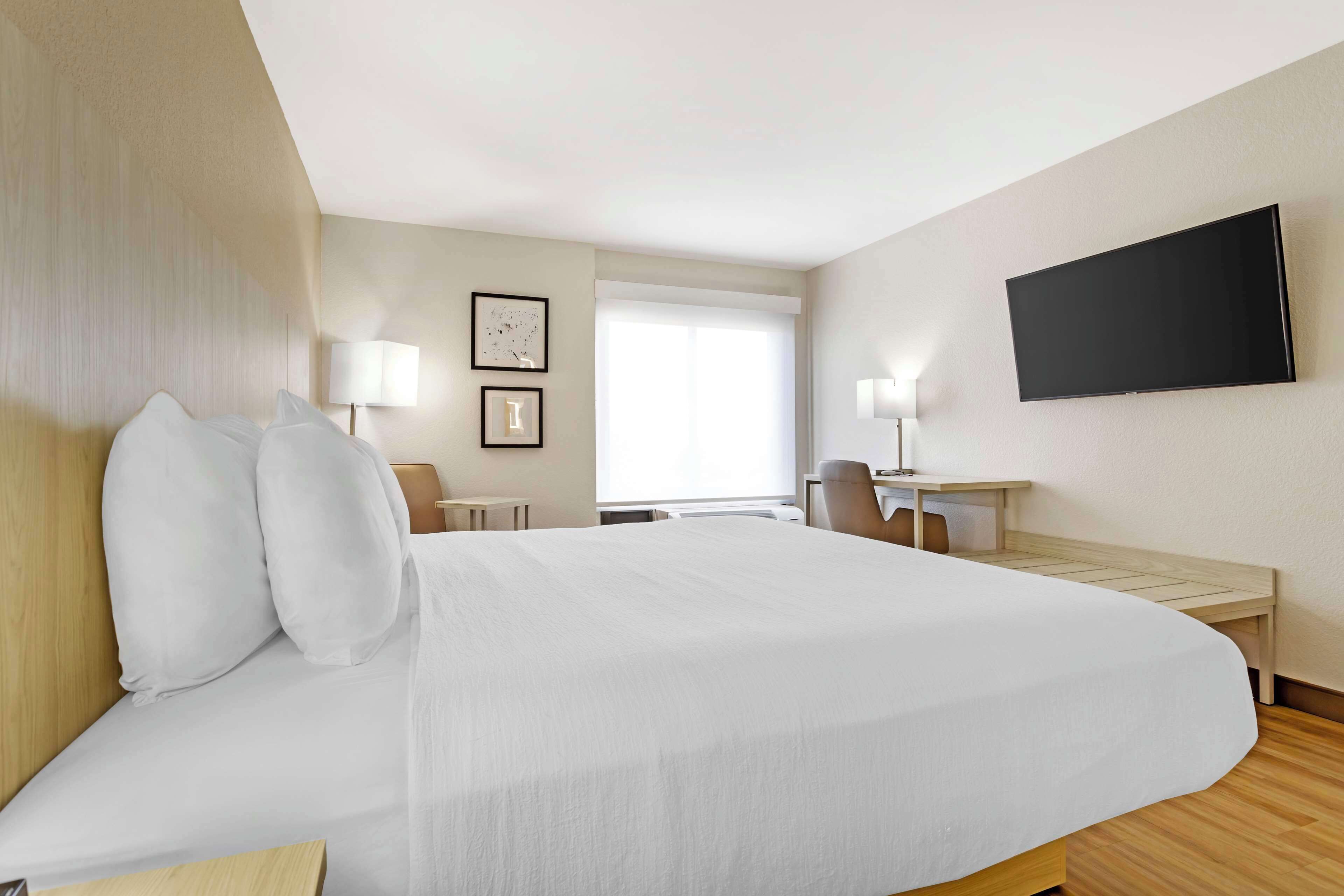 A hotel room with a large bed, a desk, a chair, a TV mounted on the wall, and a window with blinds.