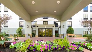 A hotel entrance with a covered walkway, a central glass door, and a landscaped garden with various plants. - Best Western Plus Sugar Land/Stafford (Sugar Land)