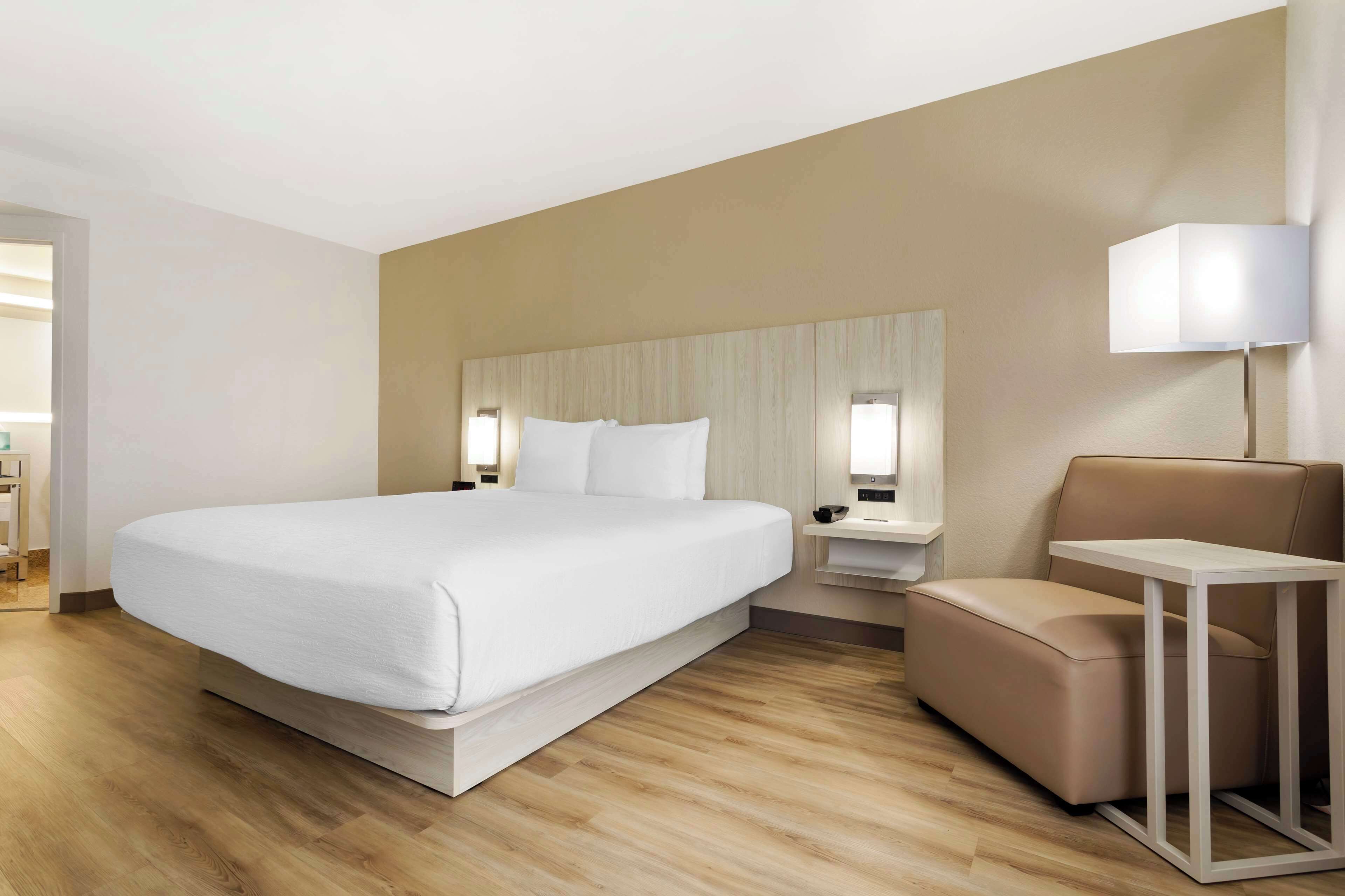 A modern hotel room with a large bed, a bedside table, a chair, and a lamp.