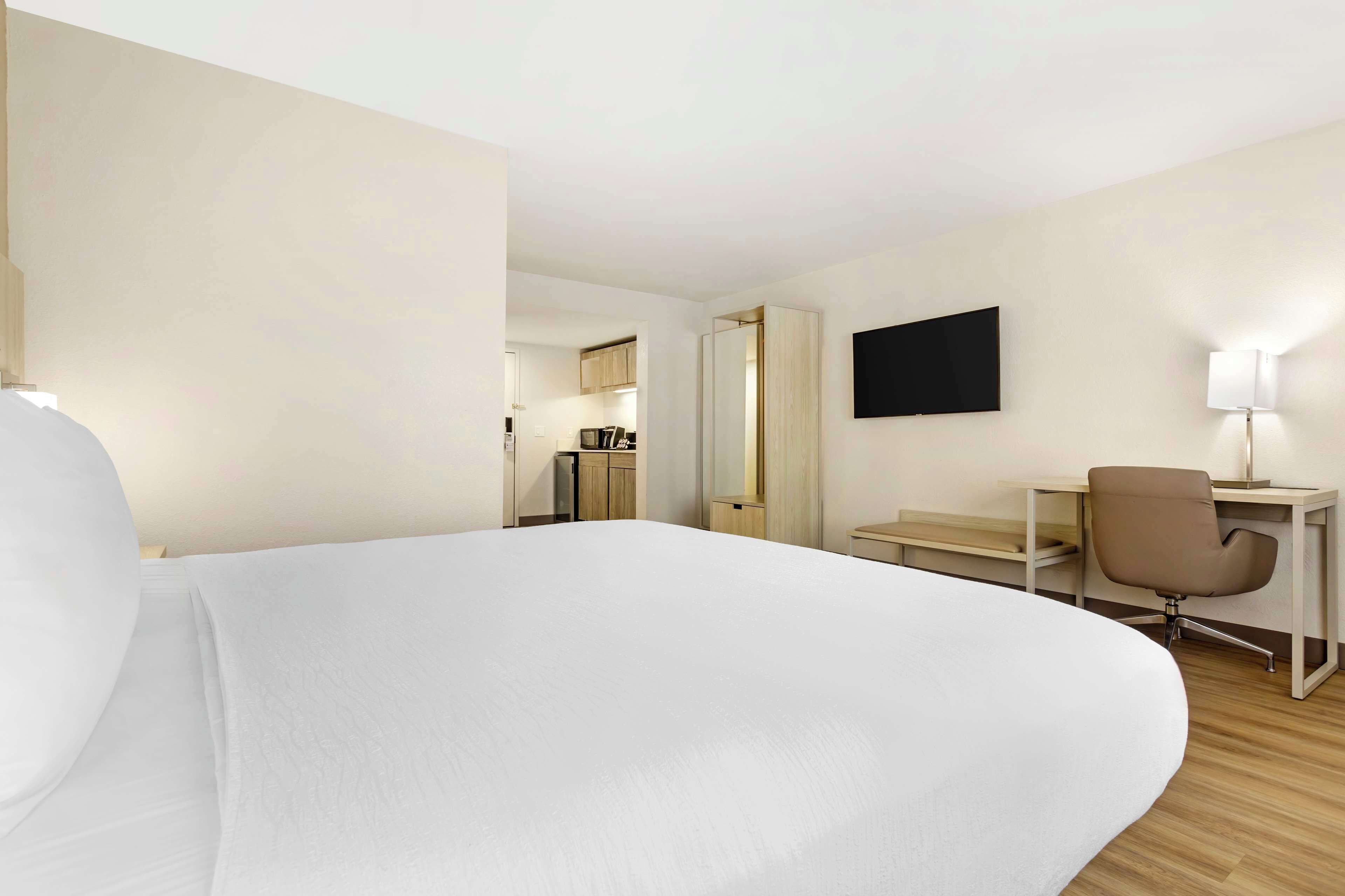 A hotel room with a large bed, a desk, a chair, a TV, and a kitchenette in the background.