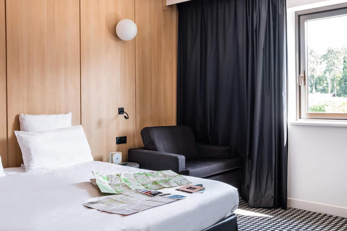 A hotel room with a bed, a desk, a chair, a window with curtains, and a wall-mounted light.